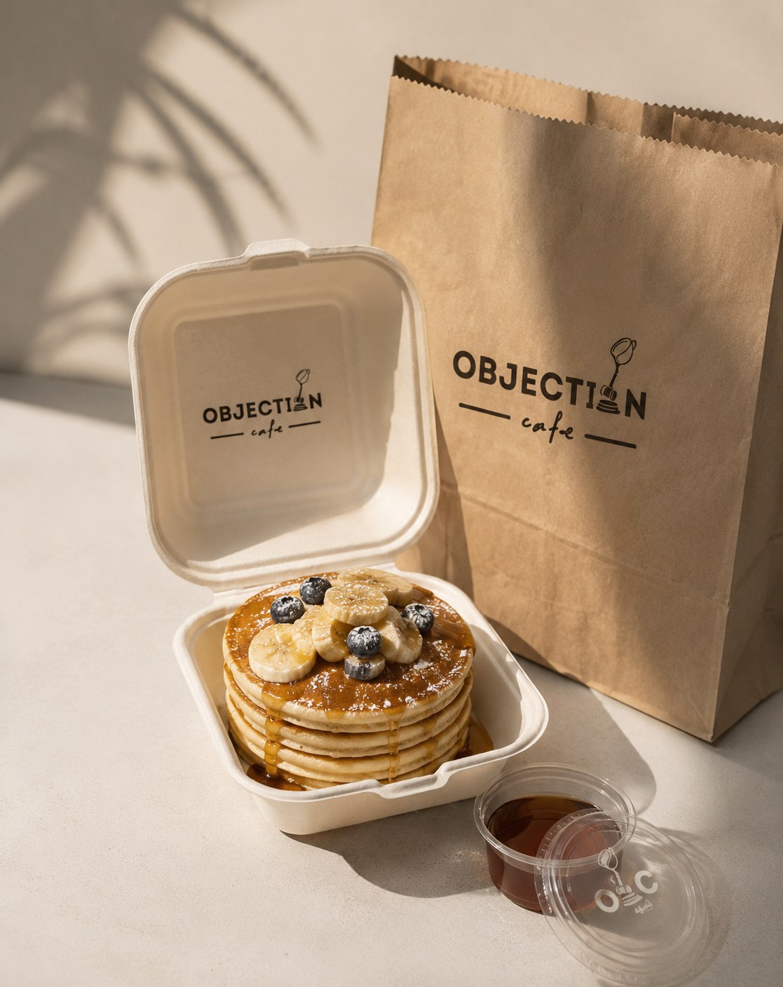 Pancakes with banana and blueberries in an Objection Cafe takeout box, next to a branded bag